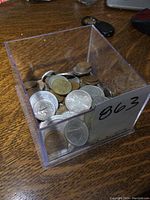 Clear plastic bin holding various vintage foreign coins scattered inside, including several silver and bronze tones.