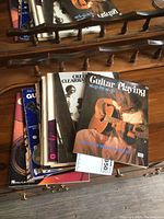 Photo showing a collection of guitar playing books stacked on a wooden surface with a partial view of a guitar stand.