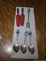 Group photo showing all items: 3 souvenir spoons with enamel designs, 2 Georg Jensen butter spreaders with red leather handles and sheath, and 1 small fork with ornate handle