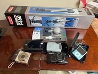 Showing the Otis T-460 tripod box, two Canon cameras, a white Canon battery charger, a blue iPod Nano with earphones in a black case, and the sealed Polaroid camera cleaning kit.