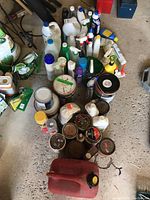 Image of many assorted opened cleaning products, paint solvents, and automotive fluid containers arranged on the concrete floor of a garage.