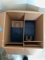 Top view of the open brown cardboard box containing two compartments filled with neatly stacked blue paper gift bags with twisted paper handles.
