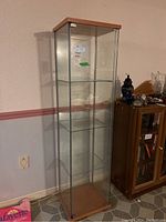 Front angled view of the tall glass display cabinet showing wooden top and bottom with three internal glass shelves, in a room corner next to another cabinet.