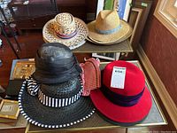 Six assorted hats on a glass table: a large black straw hat with a black and white striped band, a smaller dark straw hat with pink and grey checkered band, a black hat with sheer black fabric overlay, a red felt hat with dark purple band, a tan straw hat with blue band, and a dark grey fedora with a brownish band and buckle.