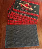 Stack of six holiday-themed mats showing teddy bear in sleigh and plaid edges with one rectangular plain weather mat in front.