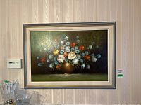 Framed oil on canvas floral still life painting hanging on wall with adjacent glassware on shelf.