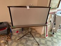 Full view of the projection screen partially raised on tripod stand, showing the screen surface and part of the surroundings.