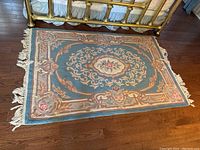 Full view of the rectangular wool rug on a wooden floor, showing blue background and floral border pattern with fringes.