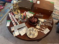 Full overhead view of all items on round wooden table including coffee flask, boxed knives, flatware sets, silver cruet set, Royal Albert utensils and bowls, Royal Worcester ramekin, and cheese board with spreaders.