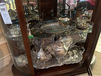 Two shelves filled with assorted silver plated tea service items including trays, teapots, condiment servers, bowls, and candlesticks shown inside a wooden and glass display case.