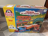 Front and side views of the Thomas The Tank Engine and Friends big set box showing the assembled train, carriages, bus, bridge, station, and tunnel.