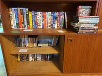 Three-shelf wooden unit with a collection of DVDs on top shelf and VHS tapes on middle and bottom shelves. The DVDs are stacked and lined up, the VHS tapes in original cases.