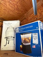 Two boxed beverage dispensers stacked in wooden barn/shed interior. The glass beverage dispenser box shows a ribbed glass jar with metal stand and lid. The plastic dispenser box is blue and white with an image of the container holding lemonade with lemon slices.