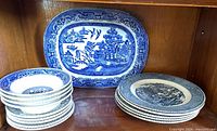 Shelf with a vintage blue and white Willow oval platter behind stacks of smaller and larger round blue and white plates with traditional patterns, displayed in wood cabinet.