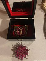 Open black decorative jewelry box showing pink multicolor butterfly brooch inside under the mirror.