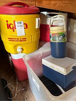 Photo showing yellow Igloo 3-gallon drinking water container with red lid and white spigot alongside several plastic coolers in red and blue.