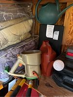 Photo of garden tools including Scotts hand spreader, watering jugs, garden shears, and cushions in a wooden shed environment.