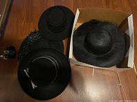Four black ladies hats displayed on wood and in box, showing one straw hat and three felt hats of varying styles and decorations.