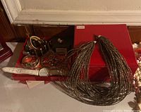 Overall view of costume jewelry including bracelets, earrings, watches, and multi-strand necklaces on and around red gift box.