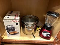 Photo of Cooks 3-in-1 grill, waffle, and sandwich maker box, two large stainless steel pots stacked with strainer lid on top, and an Oster red blender with glass jar and black lid inside a cabinet.