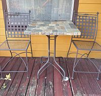 Three-piece outdoor patio set with tile table showing stone tile pattern and iron frame, two matching lattice back iron chairs on wood deck.