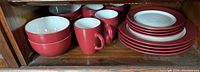 View of four red dinner plates stacked, four small sauce dishes stacked, four red mugs, and four red bowls all placed on a wooden shelf.