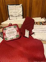 Photo showing boxed Martha Stewart Christmas guest towels, red quilted oven mitt, Christmas pot holder with plaid border and text, red woven placemat, and neatly folded linens.