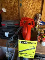 Photo showing Ryobi ONE+ red electric blower resting on its original box with cord coiled around it and black tool cases in background.