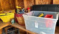 Photo of shelving with a clear plastic bin full of hand tools and a small red toolbox, a round wooden basket filled with miscellaneous items, yellow plastic container, WD-40 can, small green can, and a black plastic storage box in the background.