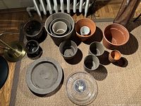 Several assorted pots and saucers including terracotta, gray plastic, and dark glazed pots in various sizes displayed on a woven floor mat.