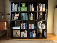 Front view showing the 16 compartments filled with books and boxes to illustrate size. Shelf is square with black-brown finish. Some wear marks visible on lower cubes.