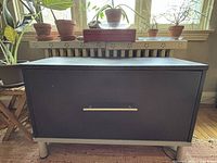 Front view of the black IKEA filing cabinet / TV stand showing flat top surface, single drawer with crooked handle, four silver legs, and visible cracks on top veneer.