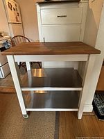 Front view of the IKEA kitchen island showing its white frame, wood top, two metal shelves, and caster wheels.