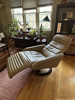 Beige leather recliner in semi-reclined position showing channel stitched seat, backrest, extended footrest and black circular swivel base.