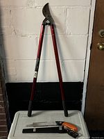 Full view of the loppers with red handles and pruning saw with orange handle placed against a white wall.