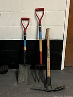 Front view of the set of three short handled gardening tools including spade, garden fork, and mattock pick seen standing on the floor against a white and black wall.