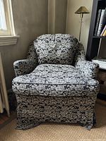 Front view showing chair covered in black fabric with white floral rose motif, cushioned armrests and seat, fabric skirt below seat, chair on casters