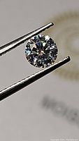 Close-up photo of one round brilliant cut moissanite held by tweezers showing its clarity and brilliance.