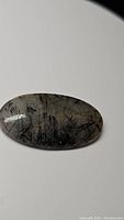 Oval cabochon black rutilated quartz with black tourmaline inclusions photographed on a white background, showing transparent quartz with needle-like black inclusions inside.