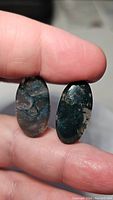 Photo showing front detail of two oval moss agate cabochons held between fingers highlighting color and inclusion patterns.