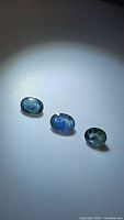 Three oval cut sapphires arranged in a row under lighting highlighting the natural blue-green multicolor and visible inclusions.