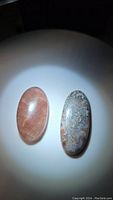 Two oval cabochon gemstones on black background spotlit: pinkish-orange translucent strawberry quartz and patterned white crazy lace agate