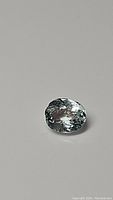 Oval natural aquamarine gemstone with visible facets and high clarity shown against a light grey background.