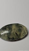 Close-up photo of a translucent oval cabochon mossy green prehnite gemstone with black rutilated and tourmaline inclusions.