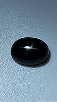 Oval cabochon black star diopside gemstone with visible star effect under light on textured light blue background.