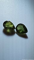 Pair of pear cut green sapphires placed on a light textured background showing color and cut details.