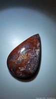 Polished red crazy lace agate pear-shaped cabochon gemstone on neutral background, showing natural lace and swirl patterns clearly.