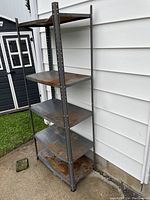 Metal shelving unit with four shelves, showing rust and wear, photographed outside next to white siding and grass.