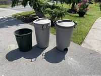 Full view of three garbage cans - two tall beige with lids and one short dark green without lid, arranged outside on pavement.