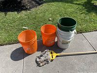 Photo of five 5-gallon buckets (three orange with Home Depot logo, one white, one green) and one mop with yellow wringer on concrete surface outdoors, grass background
